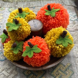 May include: A selection of handmade pom-pom pumpkins in yellow and orange, with green felt leaves and brown stems. The pumpkins are arranged on a brown plate, ideal for autumn decorations.