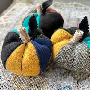 May include: Three stuffed fabric pumpkins featuring patchwork panels in yellow, navy blue, dark green, brown, and teal. Each pumpkin has a textured, light-coloured wrapped stem and a small, pointed black fabric leaf attached at the top.