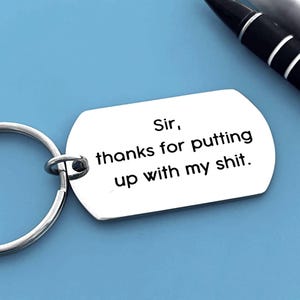 May include: A silver metal keyring with a tag that reads "Sir, thanks for putting up with my shit." The tag is a rounded rectangle. The keyring is on a blue surface, with pens visible in the background.