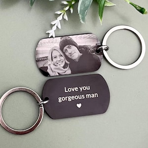 May include: Two rectangular metal keychains with rounded corners and silver-toned split rings. One keychain features a black and white engraved photograph of two people. The other keychain is black with the white engraved text Love you gorgeous man.