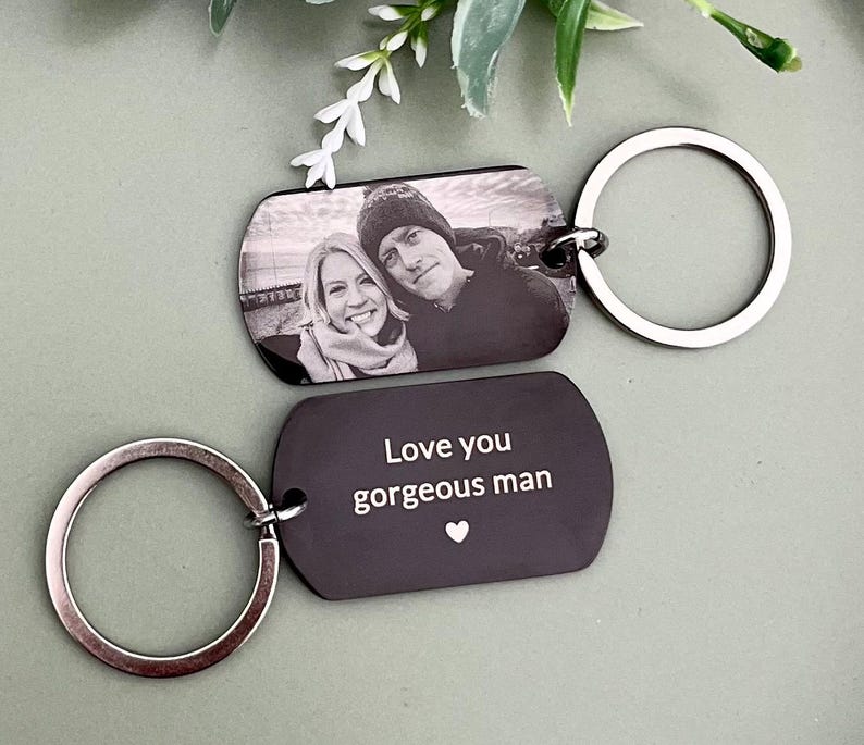May include: Two rectangular metal keychains with rounded corners and silver-toned split rings. One keychain features a black and white engraved photograph of two people. The other keychain is black with the white engraved text Love you gorgeous man.