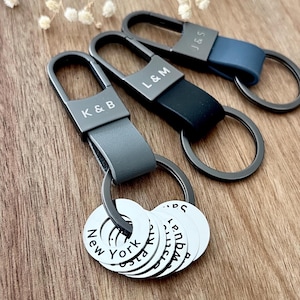 Puede incluir: Tres llaveros personalizados con iniciales y nombres de ciudades. Los llaveros tienen un cierre metálico y una correa de cuero en gris, negro y azul. Cada llavero tiene un anillo con etiquetas circulares blancas grabadas con nombres de ciudades.