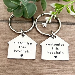 May include: Two silver keychains with house-shaped pendants. Each pendant has the text "customise this keychain" and a small heart symbol. The keychains are on a wooden surface, with greenery in the background.