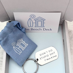 May include: A silver keychain with the text "Don't do stupid shit" and a heart symbol followed by "Mum & Dad". The keychain is in a small blue gift bag with a white beach hut and surfboard design. The bag is in a white box with a white card that has a beach hut and surfboard design and the text "The Beach Deck".