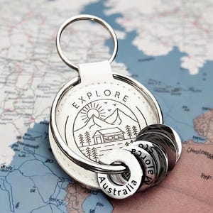 Puede incluir: Un llavero circular de cuero blanco con una ilustración de líneas negras de una cabaña, montañas y el sol, con la palabra EXPLORE impresa encima. Se adjuntan varios anillos de metal plateado, uno de ellos grabado con la palabra Australia.