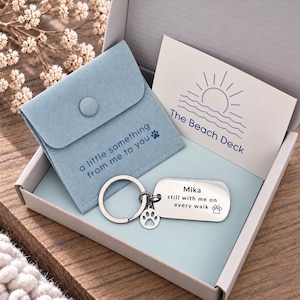 Puede incluir: Un llavero rectangular plateado con un peque&ntilde;o dije de huella, grabado con el texto 'Mika still with me on every walk'. Se presenta en una caja con una bolsa azul claro etiquetada 'a little something from me to you' y una tarjeta que dice 'The Beach Deck'.