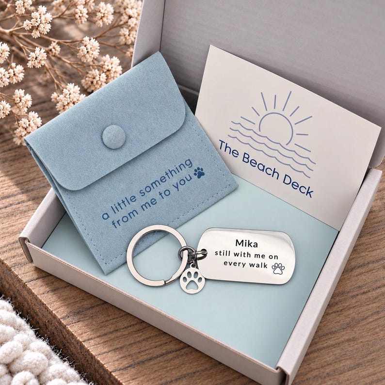 Puede incluir: Un llavero rectangular plateado con un peque&ntilde;o dije de huella, grabado con el texto 'Mika still with me on every walk'. Se presenta en una caja con una bolsa azul claro etiquetada 'a little something from me to you' y una tarjeta que dice 'The Beach Deck'.