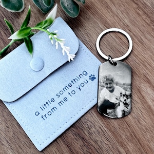 May include: A silver-toned metal dog tag keychain featuring a black and white engraved photograph of a person and a dog. It is accompanied by a light blue fabric pouch with the text "a little something from me to you" and a paw print icon.