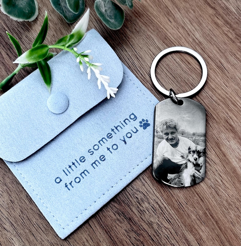 May include: A silver-toned metal dog tag keychain featuring a black and white engraved photograph of a person and a dog. It is accompanied by a light blue fabric pouch with the text "a little something from me to you" and a paw print icon.