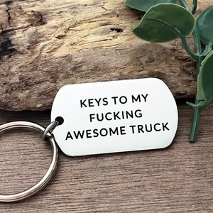 Op de afbeelding: Zilveren sleutelhanger met een zwart label waarop "Keys to my fucking awesome truck" staat.