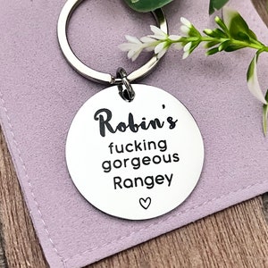 Könnte beinhalten: Ein silberner Schlüsselanhänger mit einem runden Anhänger, auf dem "Robin's fucking gorgeous Rangey" mit einem kleinen Herzsymbol steht.