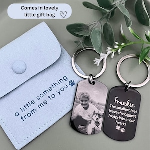 May include: Two rectangular metal keychains with silver-toned rings. One keychain features a black and white photograph of a person and a dog. The other keychain is black with the text Frankie, The smallest feet leave the biggest footprints in our hearts, and two paw prints. A light blue fabric pouch with the text a little something from me to you and a paw print is included.