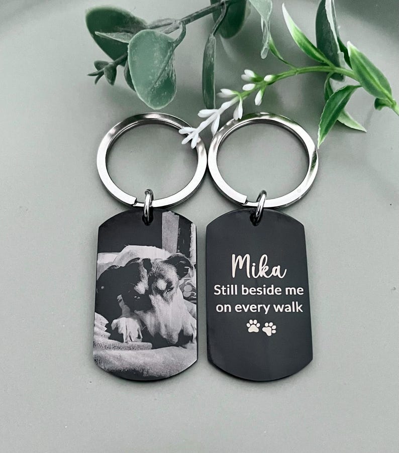 May include: Two black metal dog tag keychains with silver split rings. One tag features a laser-engraved black and white photograph of a dog. The other tag displays the white text Mika, Still beside me on every walk, and two paw print icons.