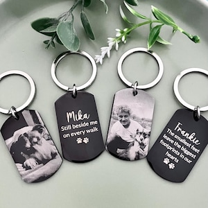 May include: Four rectangular black metal keychains with silver split rings. Two keychains feature engraved black and white photographs of dogs and people. Two keychains feature white text: Mika Still beside me on every walk and Frankie The smallest feet leave the biggest footprints in our hearts, both with paw print icons.