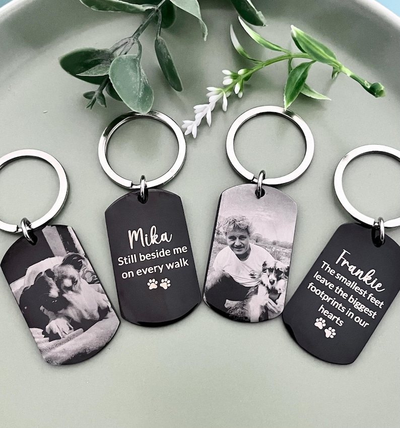 May include: Four rectangular black metal keychains with silver split rings. Two keychains feature engraved black and white photographs of dogs and people. Two keychains feature white text: Mika Still beside me on every walk and Frankie The smallest feet leave the biggest footprints in our hearts, both with paw print icons.