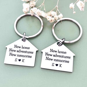 May include: Two silver-toned metal keyrings shaped like houses. Each keyring features the engraved text: New home, New adventures, New memories, and the letters S and K separated by a heart symbol. The keyrings include circular metal rings.