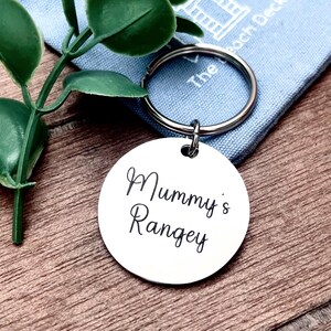 Puede incluir: Un llavero de plata con una etiqueta redonda que dice "Mummy's Rangey" en escritura cursiva.