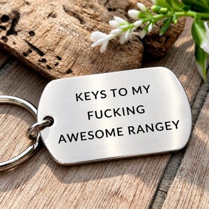 May include: A silver-toned metal dog tag keychain with a brushed finish and a circular split ring. The tag is engraved with the black text: KEYS TO MY FUCKING AWESOME RANGEY. The keychain is positioned on a textured wooden surface with a small plant.