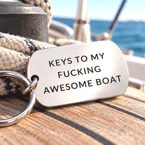 Könnte beinhalten: Ein silberfarbener Metall-Schlüsselanhänger in Form einer Erkennungsmarke mit einem runden Spaltring. Die Marke ist mit dem schwarzen Text graviert: KEYS TO MY FUCKING AWESOME BOAT. Der Schlüsselanhänger liegt auf einem dicken weißen Seil und einem Holzdeck.