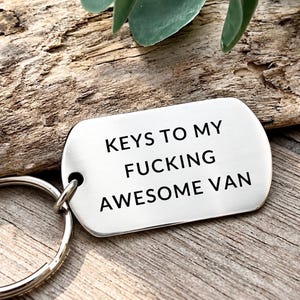May include: A silver-coloured metal dog tag keyring with rounded corners and a circular split ring. The surface is engraved with the black text: KEYS TO MY FUCKING AWESOME VAN. The tag has a smooth, polished finish and a single hole for the ring attachment.