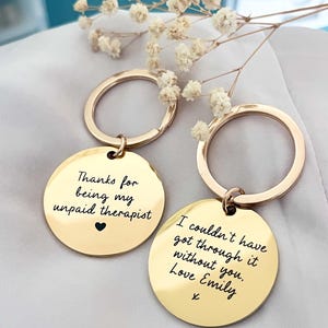 Può includere: Due portachiavi color oro con ciondoli rotondi. Un portachiavi recita "Thanks for being my unpaid therapist" con un cuore. L'altro dice "I couldn't have got through it without you. Love Emily".