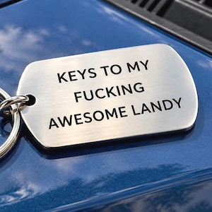 Könnte beinhalten: Ein rechteckiger Schlüsselanhänger aus gebürstetem, silberfarbenem Metall mit abgerundeten Ecken und einem Spaltring. Der Anhänger trägt den schwarzen Gravurtext: KEYS TO MY FUCKING AWESOME LANDY. Die Metalloberfläche hat eine horizontale Maserung.