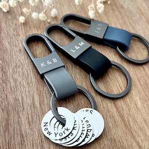 May include: Three metal keychains with matte black carabiners and leather loops in grey, black, and navy blue. Each carabiner is engraved with initials K & B, L & M, or J & S. Attached to the rings are multiple silver circular charms with text.
