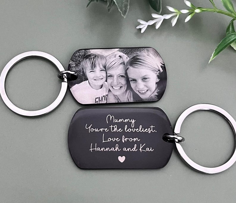 May include: Two rectangular metal keychains with rounded corners and silver-toned split rings. One keychain features a black and white photograph of three people. The other is black with white engraved text: Mummy You're the loveliest. Love from Hannah and Kai, and a small pink heart.
