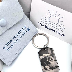 May include: A silver-toned metal dog tag keychain featuring a black and white photograph of a dog. The keychain is accompanied by a light gray fabric pouch with the text "a little something from me to you" and a paw print, plus a white card.