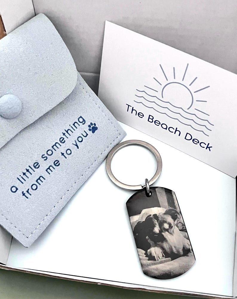 May include: A silver-toned metal dog tag keychain featuring a black and white photograph of a dog. The keychain is accompanied by a light gray fabric pouch with the text "a little something from me to you" and a paw print, plus a white card.