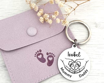 Pode incluir: Um porta-chaves prateado com o nome "Isobel" gravado num pendente redondo, com um design em forma de coração com pegadas de bebé e as palavras "Mummy" e "Daddy". Uma bolsa lilás com pegadas de bebé está no fundo.