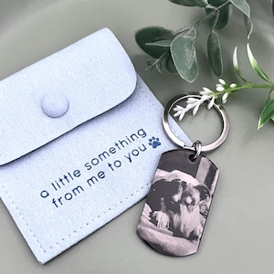 May include: A light blue suede-like pouch with a snap closure featuring the text 'a little something from me to you' and a blue paw print icon, paired with a silver-toned metal dog tag keychain displaying a black and white photo of a dog.