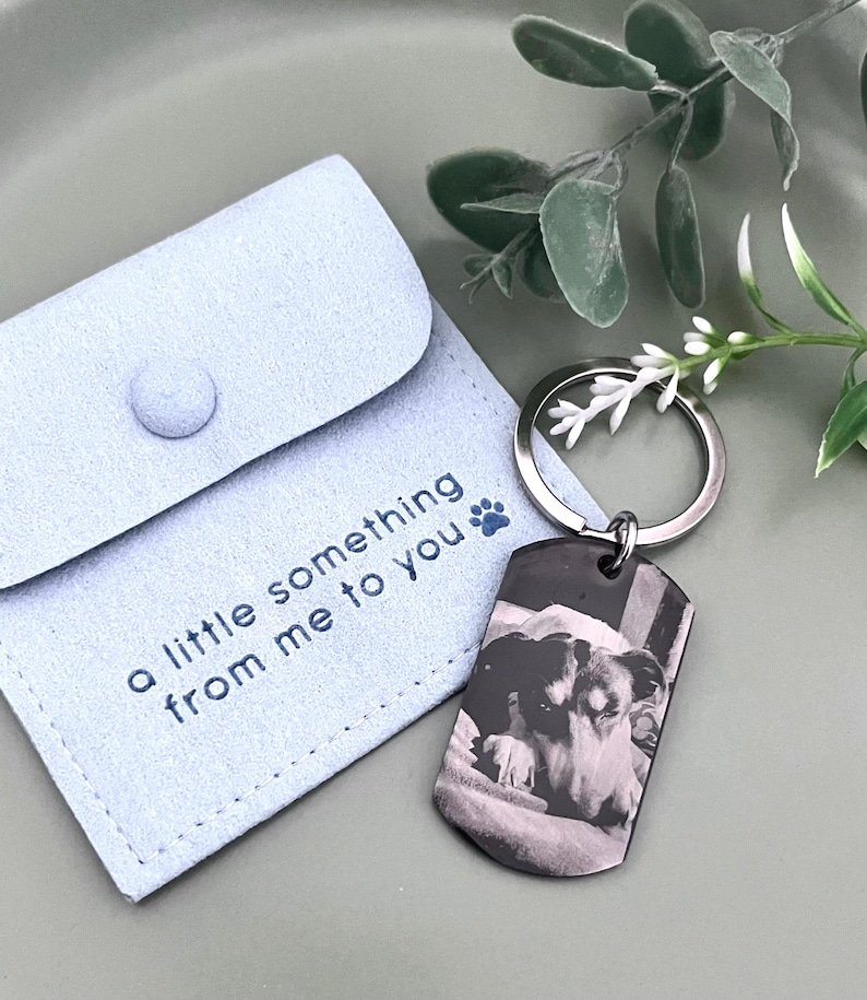 May include: A light blue suede-like pouch with a snap closure featuring the text 'a little something from me to you' and a blue paw print icon, paired with a silver-toned metal dog tag keychain displaying a black and white photo of a dog.
