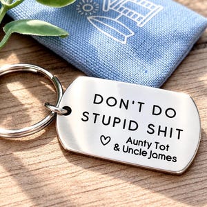 Puede incluir: Una etiqueta rectangular de metal plateado para llavero con esquinas redondeadas y una anilla. El frente está grabado con el texto DON'T DO STUPID SHIT seguido de un símbolo de corazón y los nombres Aunty Tot & Uncle James en negro.