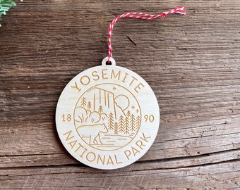 May include: A round wooden ornament with a laser-engraved design of Yosemite National Park. The design includes a bear, trees, a mountain, and the text "Yosemite National Park 1890".