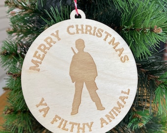 Może przedstawiać: Drewniana ozdoba choinkowa z napisem "Merry Christmas Ya Filthy Animal" i sylwetką osoby trzymającej broń.