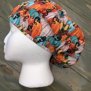 May include: A scrub cap featuring a vibrant autumnal pattern of orange, teal, and white pumpkins, green leaves, and small red berries. The fabric has a gathered design with a wide band across the front and a rounded, voluminous back section.