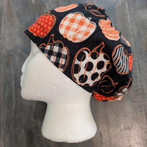 May include: A scrub cap featuring a black background with a repeating pattern of pumpkins in various styles, including orange polka dots, orange and white gingham, black and white buffalo check, and black and white stripes, worn on a white mannequin head.