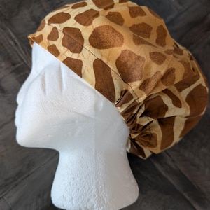 May include: A brown and tan giraffe print scrub cap. The cap is made of fabric and has a gathered elastic back. The cap is designed to cover the hair and is shown on a white mannequin head.