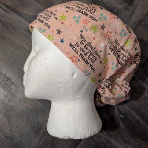 May include: A peach-colored scrub cap with a repeating pattern of colorful stars and the text "You don't have to be crazy to work here. We'll train you." The cap is designed to be worn in a medical or professional setting.