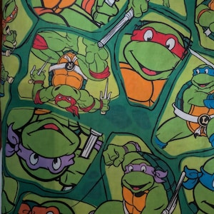 Può includere: Un tessuto verde con un motivo ripetitivo delle Tartarughe Ninja. Il design mostra diverse tartarughe che indossano maschere arancioni, rosse, viola e blu, armate di nunchaku e sai, su uno sfondo di colore verde.