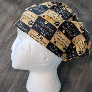 Puede incluir: Un gorro quirúrgico con estampado negro y marrón con las palabras "Mischief Managed" y huellas de patas. El gorro también tiene el texto "I solemnly swear that I am up to no good". El gorro está sobre una cabeza de maniquí blanca.