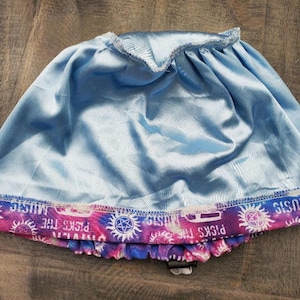 May include: A light blue satin skirt with a purple, pink, and white tie-dye trim featuring the text "Picks The Music" and a symbol.