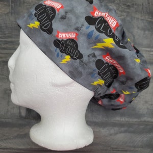 May include: A gray scrub cap with a pattern of black clouds, yellow lightning bolts, and blue raindrops. The words "CERTIFIED BLACK CLOUD" are printed on red banners. The cap is displayed on a white mannequin head.