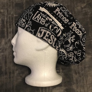 May include: A black and white patterned scrub cap with religious text including "Lamb of God" and "Prince of Peace". The cap is displayed on a white mannequin head, showcasing the design and fit. The cap is made of fabric.