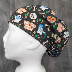May include: Black scrub hat with a Halloween theme. The hat features a variety of spooky designs, including ghosts, pumpkins, bones, and stars. The text "Boo-Boo Crew" is printed on the hat.