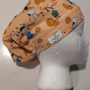 May include: A peach-colored scrub cap featuring a repeating pattern of Snoopy, Charlie Brown, and Lucy characters among pumpkins and autumn leaves. A small white fabric label on the side displays the text Caroline Dreams by Jen.