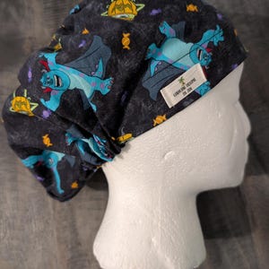 May include: A black scrub cap featuring a repeating pattern of blue monsters with bat wings, yellow pumpkins, and orange candies. A small white fabric tag is sewn onto the side with the text "Caroline Dreams by Jen" and a palm tree icon.
