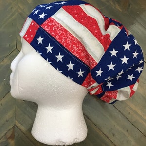 May include: A patriotic scrub cap featuring a patchwork design of the American flag. The cap displays blue panels with white stars, red stripes, and floral patterns. The cap is displayed on a white mannequin head.