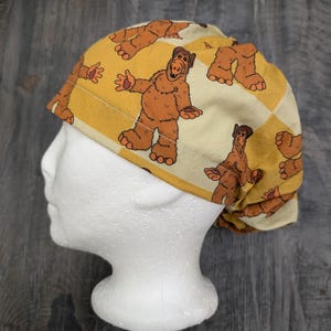 May include: A yellow and brown patterned surgical scrub hat with a cartoon character of a furry creature with a brown body and a brown head with a white face. The character has a brown nose and a brown mouth. The character is wearing a brown hat with a white brim. The hat is on a white mannequin head.